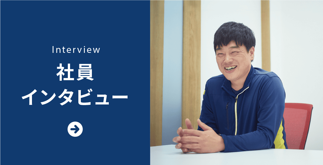 インタビュー中の社員。青いジャケットを着ており、黄色のファスナーが目立ちます。手を組んでテーブルに座っています。背景には木製の枠と赤い椅子があります。左側には青い背景に白いテキストで「INTERVIEW 社員 インタビュー」と書かれ、下に右向きの白い矢印があります。