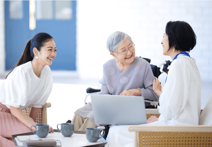 室内でテーブルを囲んで会話している3人の人物が写っています。彼らの前にはラップトップと3つのコーヒーカップがあります。1人は車椅子に乗っている高齢者のように見え、他の2人は若く見えます。3人は高齢者に向かって親しみやすく話しています。この場所はカジュアルまたはセミフォーマルな環境のようです。場面は介護、相談、または家族の集まりに関連した会話を示唆しています。